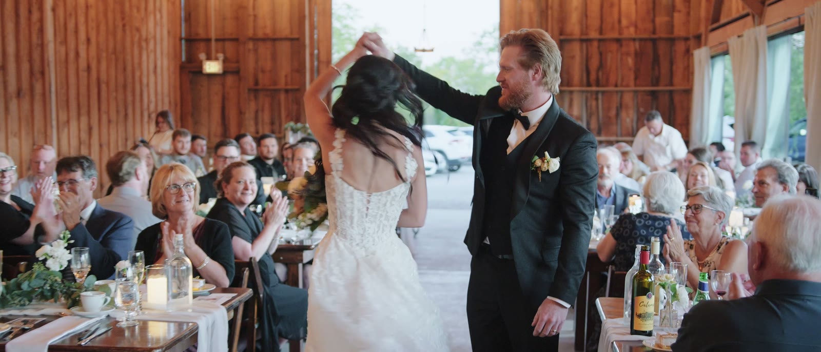 A bride and groom dancing between long guest tables inside the wooden barn, daylight coming through the tall windows along both walls.