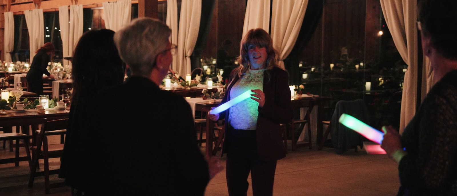 Guests standing between candlelit tables in the barn after dark, drapery and warm low light, the room much dimmer than earlier in the evening.