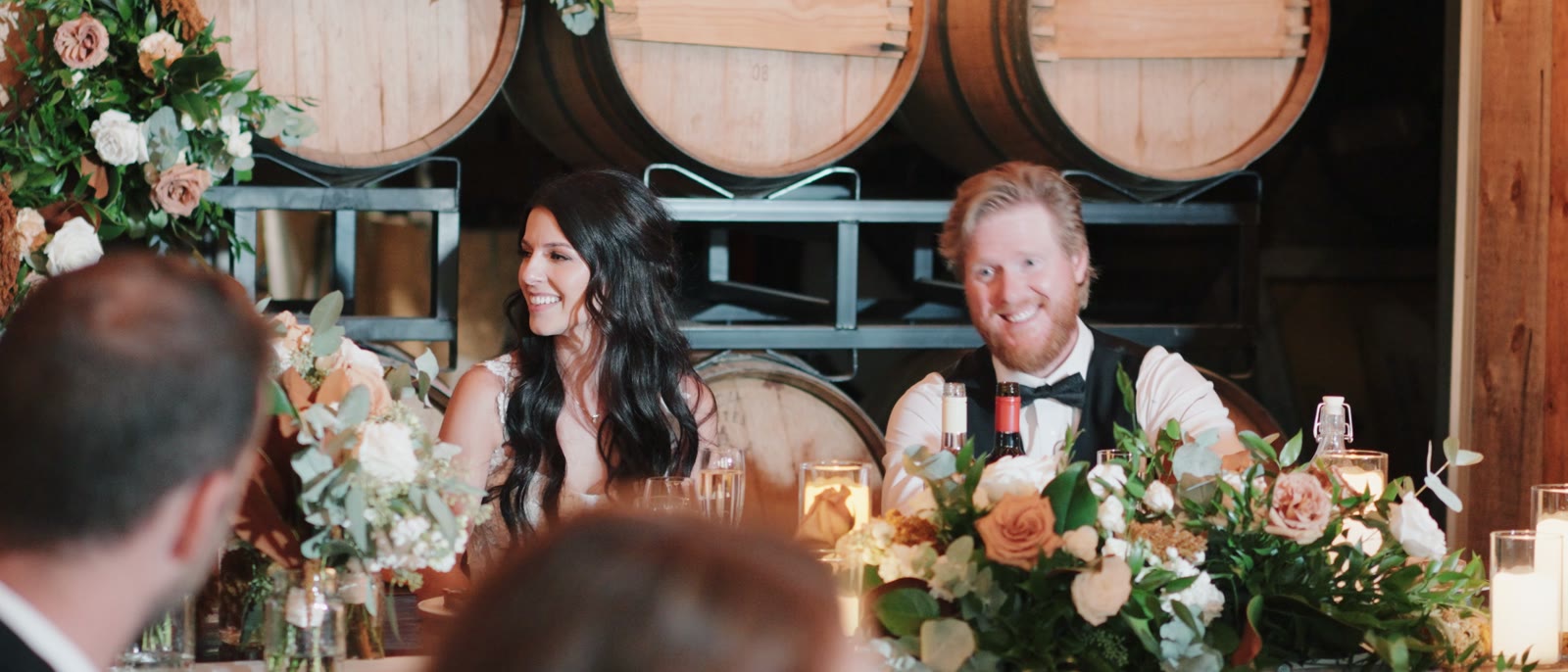 A bride and groom seated at their head table in front of three large oak wine barrels, with cream and copper floral arrangements and candles along the table.