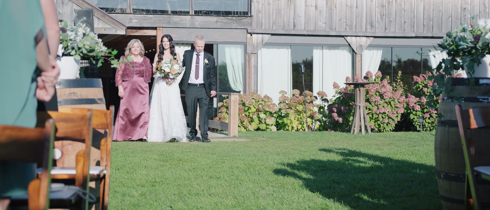A bride walking down a grass aisle with her parents toward a floral arch, with the weathered wood barn, its covered porch and hydrangeas along the edge of the lawn.
