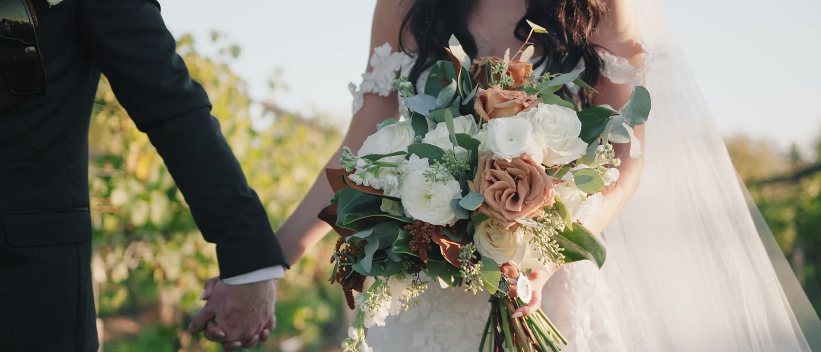 A bride and groom holding hands, her bouquet of cream roses and copper foliage in the foreground, with sunlit vineyard rows behind them.
