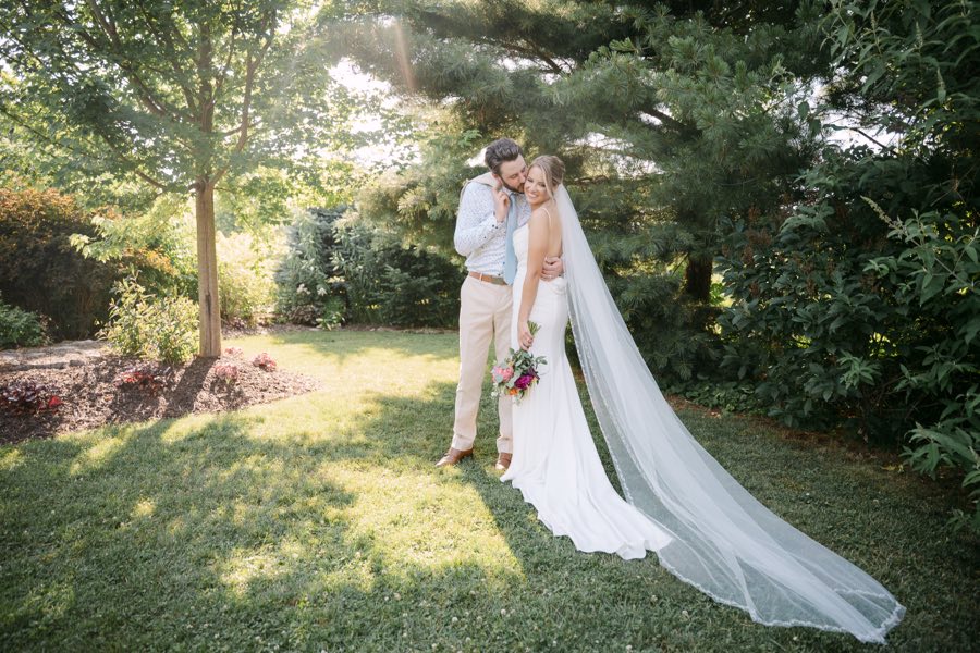 A groom kissing his bride&rsquo;s cheek in a sunlit garden