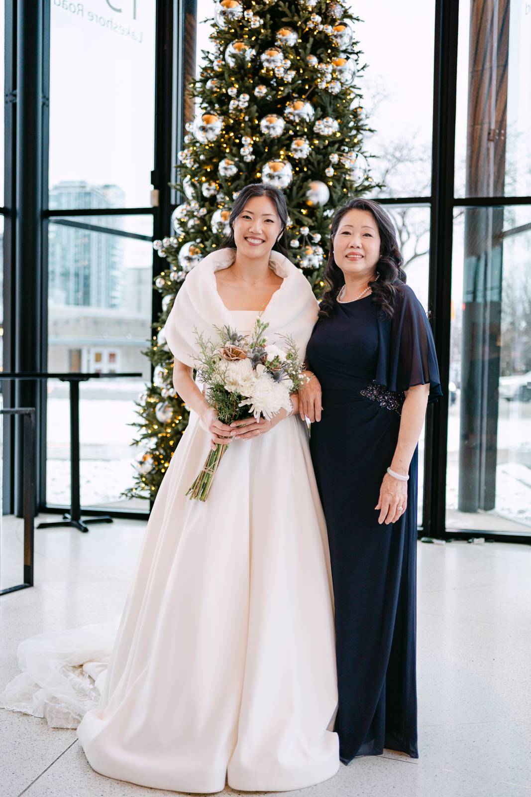 A bride in an ivory ballgown with a soft wrap over her shoulders stands beside her mother in a navy gown, in front of a tall decorated tree at the glass entrance, with snow visible on the street outside.