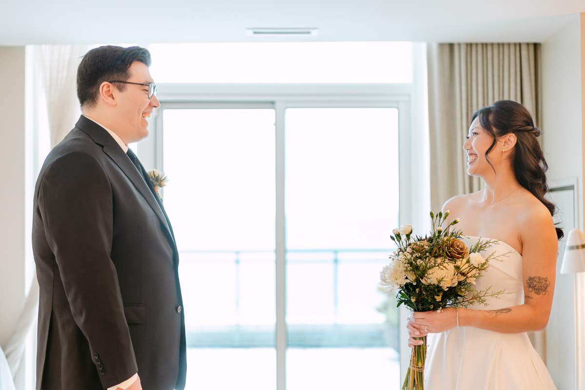 A groom in a dark suit and a bride in a strapless ivory gown holding a bouquet of cream roses and cedar, laughing at each other beside a bright balcony door.