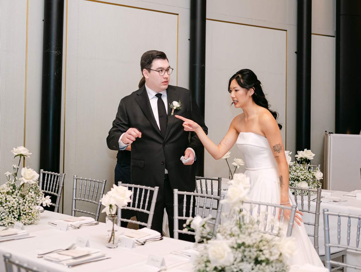 A bride and groom talking animatedly beside a long reception table set with white roses, baby's breath, silver chiavari chairs and folded napkins.