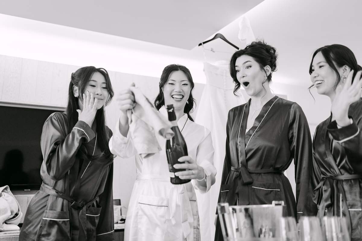 Black and white photograph of a bride in a white robe opening a bottle of sparkling wine while three bridesmaids in satin robes react with delight.