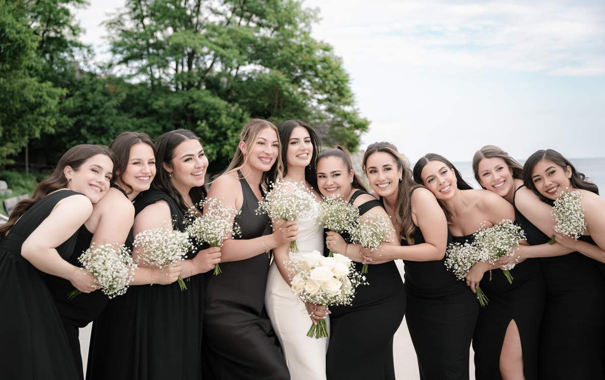 A bride holding white roses stands at the centre of ten bridesmaids in black gowns, all leaning in towards her and holding baby's breath bouquets, with trees and Lake Ontario behind them.