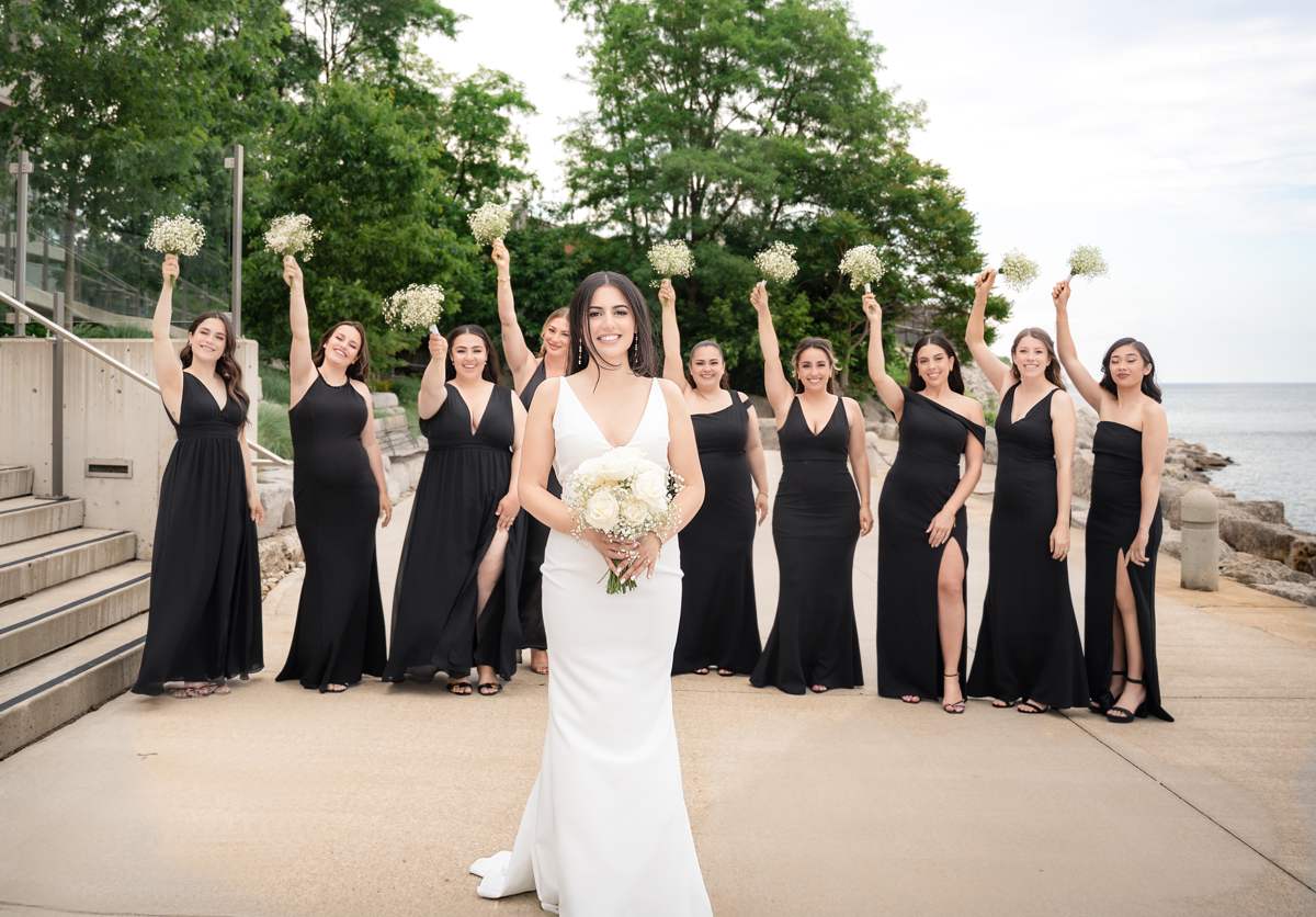 A bride holding a bouquet of white roses stands in front of ten bridesmaids in black gowns raising baby's breath bouquets, beside a concrete staircase with glass railings and the lake on the right.